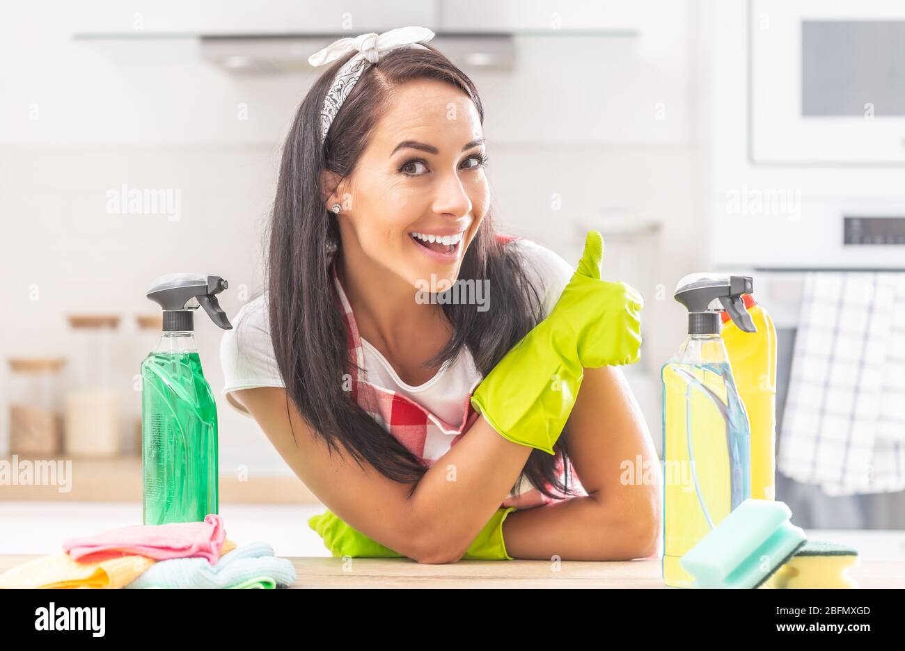 La governante si appoggia sui gomiti su una scrivania, con guanti di gomma gialla che gesturano il pollice con varie bottiglie di disinfettanti intorno a lei. Foto Stock