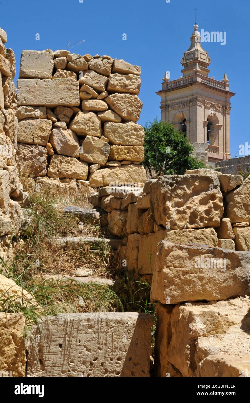 La torre della Cattedrale dell'Assunzione sorge dietro le rovine storiche della cittadella di Victoria, la capitale dell'isola di Gozo a Malta. Foto Stock