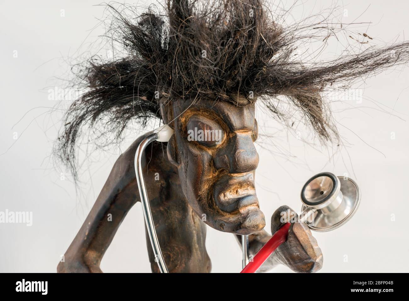 Figura intagliata in legno di uomo primitivo con gonna di paglia e lunghi capelli neri a forma di tussia, con stetoscopio. Concetto; aspirazione, ambizione, mobilità sociale. Foto Stock