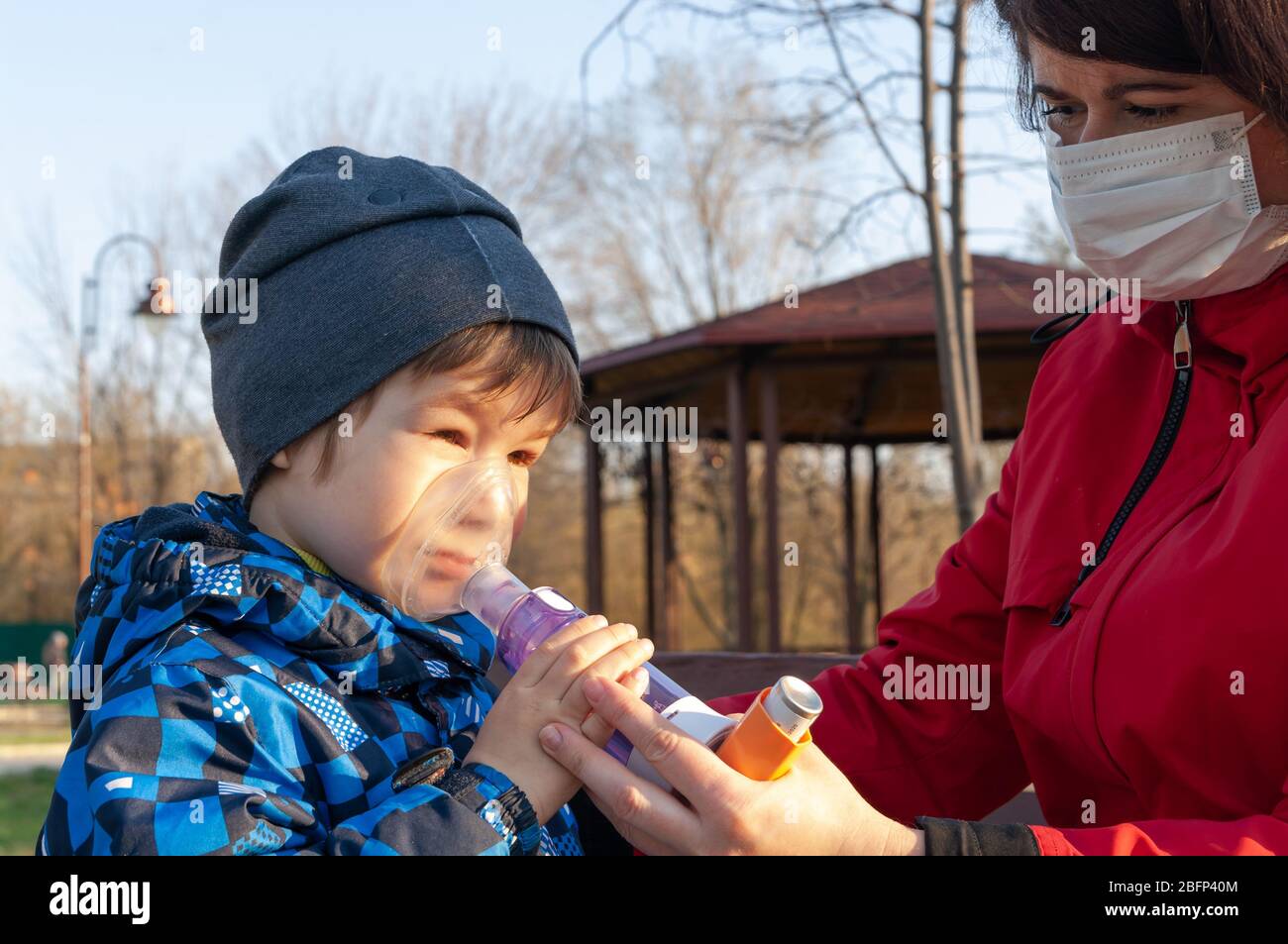 Un ragazzino affluso di asma bronchiale che riceve il trattamento con l'inalatore aerosol dalla sua mamma all'aperto Foto Stock