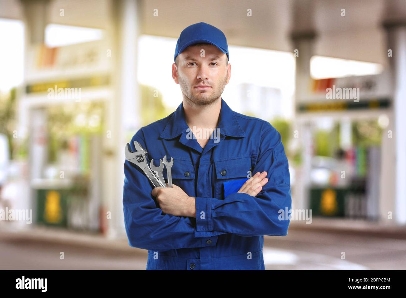 Giovane meccanico in uniforme con braccia incrociate e chiavi in piedi su una stazione di benzina sfocata Foto Stock