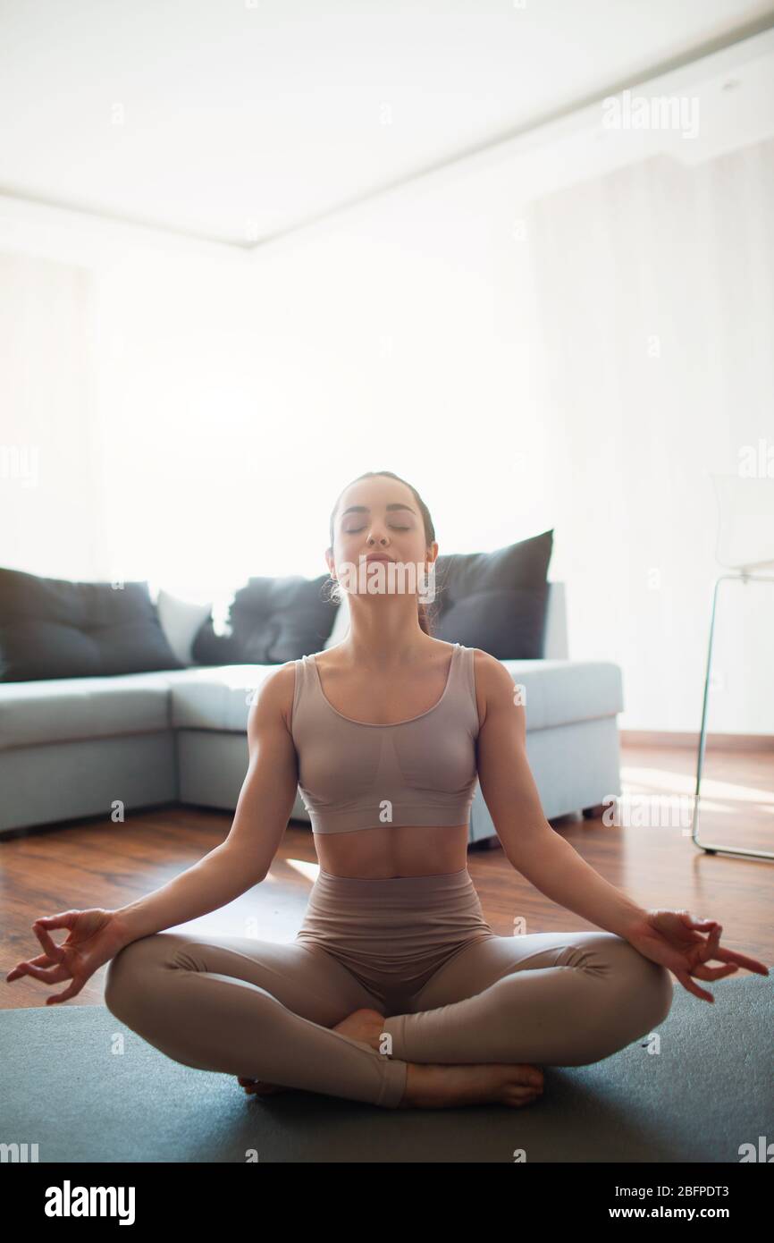 Giovane donna che fa l'allenamento di yoga in camera durante la quarantena. Ragazza siede in posizione lotus e medita con occhi chiusi. Stretching e l'esercizio in casa. Foto Stock