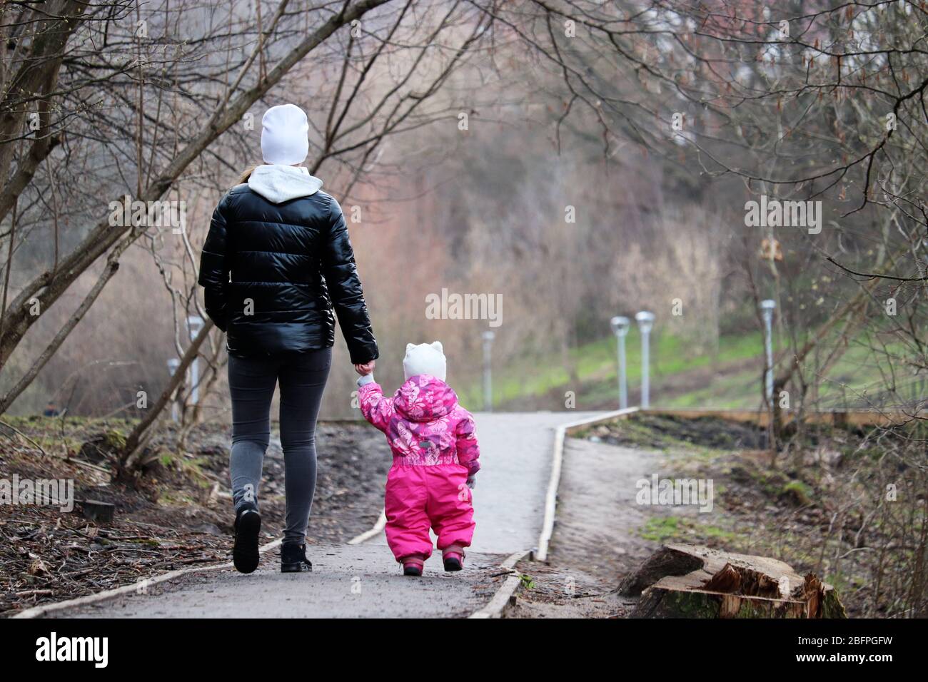 Mamma e bambino stanno camminando lungo il sentiero tenendo le mani. Giovane donna snella guida bambina in un parco primaverile, concetto per la maternità, madre singola Foto Stock