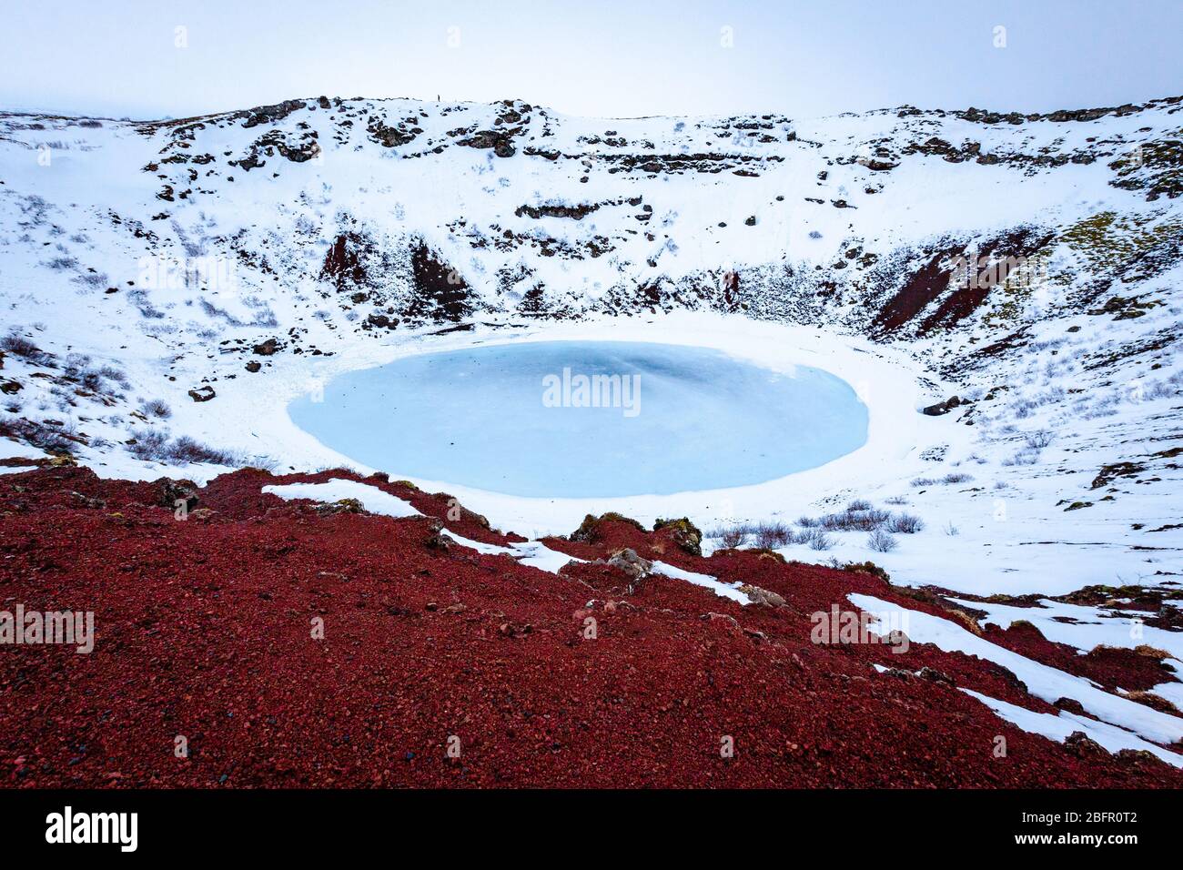 Lago vulcanico Kerid Crater in Islanda, ghiacciato blu in una giornata innevata in inverno con roccia vulcanica rossa in primo piano Foto Stock