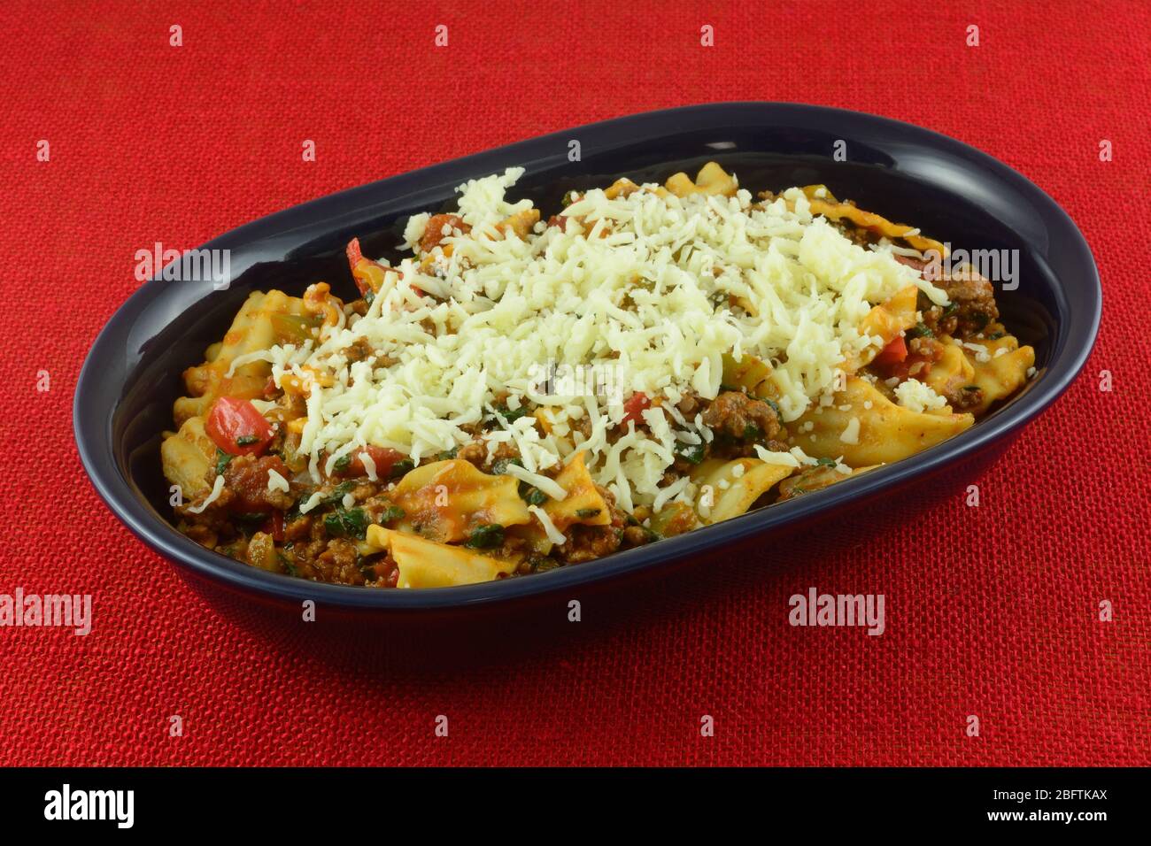 Casseruola di lasagne con carne di hamburger, spinaci, pomodori, cipolla e peperoni verdi affumicati con formaggio grattugiato in una ciotola blu su rosso Foto Stock
