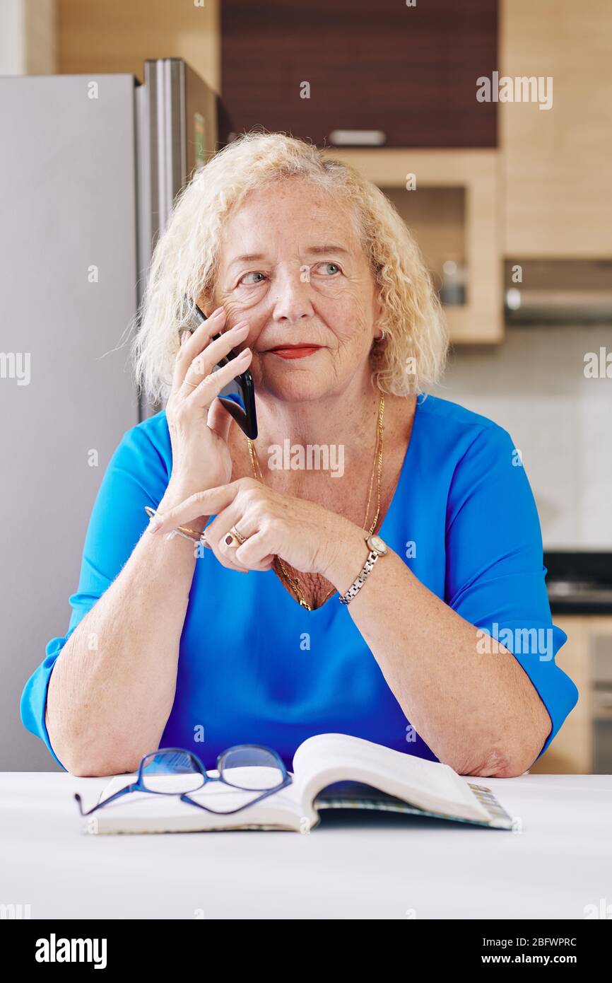 Donna anziana seduta al tavolo da cucina con libro aperto e conversazione al telefono Foto Stock