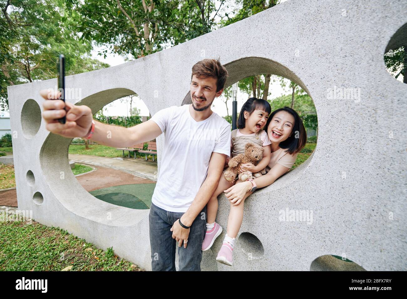 Sorridente bel giovane uomo che prende selfie con la sua moglie e figlia piccola prett quando stanno trascorrendo il tempo che parcheggio Foto Stock
