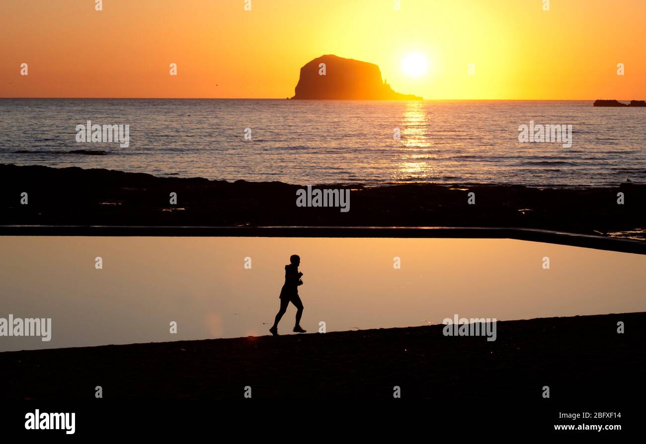 North Berwick, Bass Rock, Sunrise, East Lothian, Scozia, Regno Unito. 20 aprile 2020. Splendida scena subito dopo l'alba, quando il sole apparve da dietro il Bass Rock, Regno Unito. Temperatura 3 gradi poco o nessun vento. Un po' di calma mentre la minaccia del Coronavirus incombe ancora. Nella foto: jogger che prende il loro excercise permesso che corre oltre la piscina perfettamente ancora remare. Berwick nord mare Foto Stock