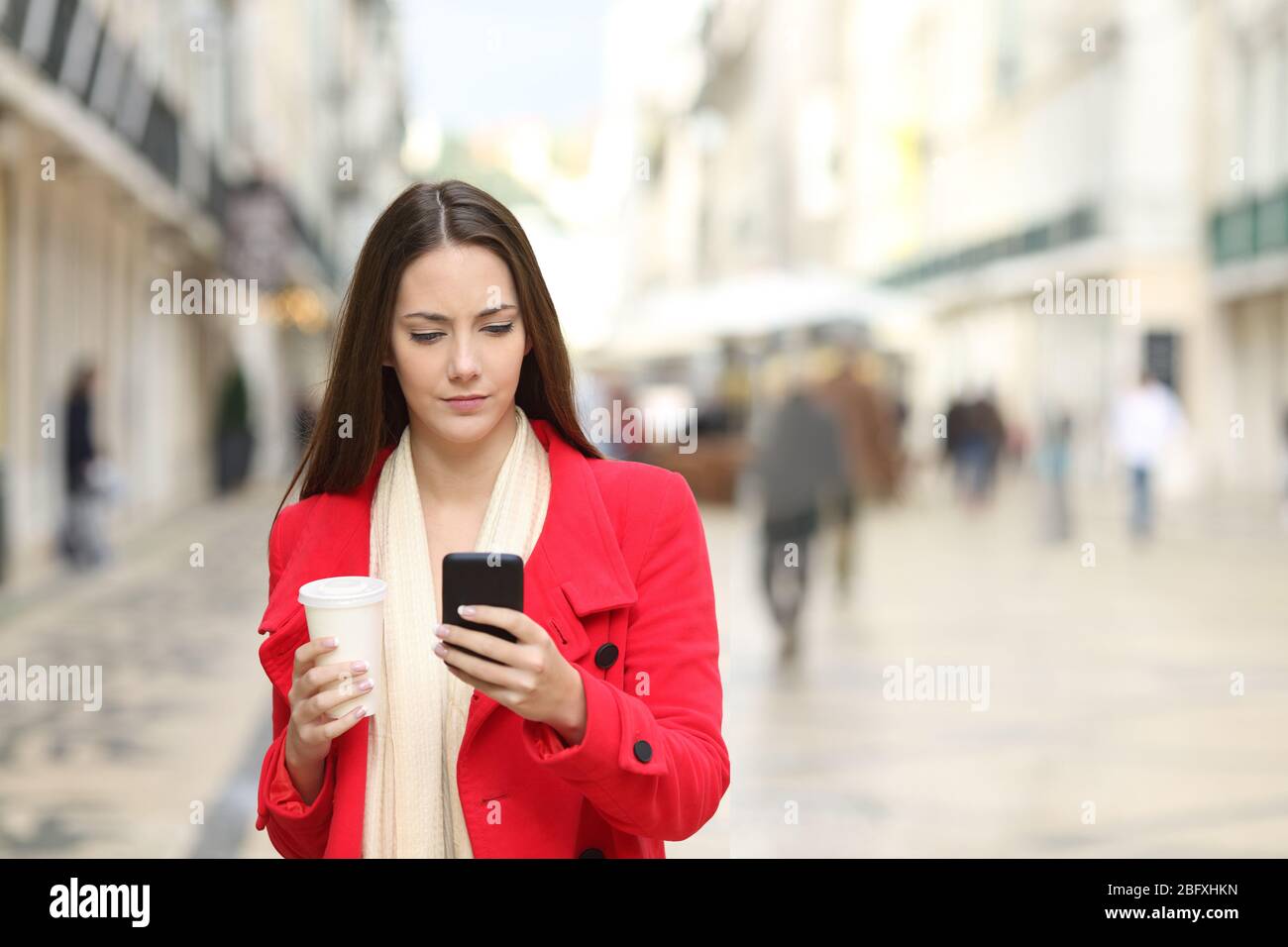 Vista frontale di una donna seria in cappotto rosso con smartphone con caffè da asporto che cammina per strada in inverno Foto Stock