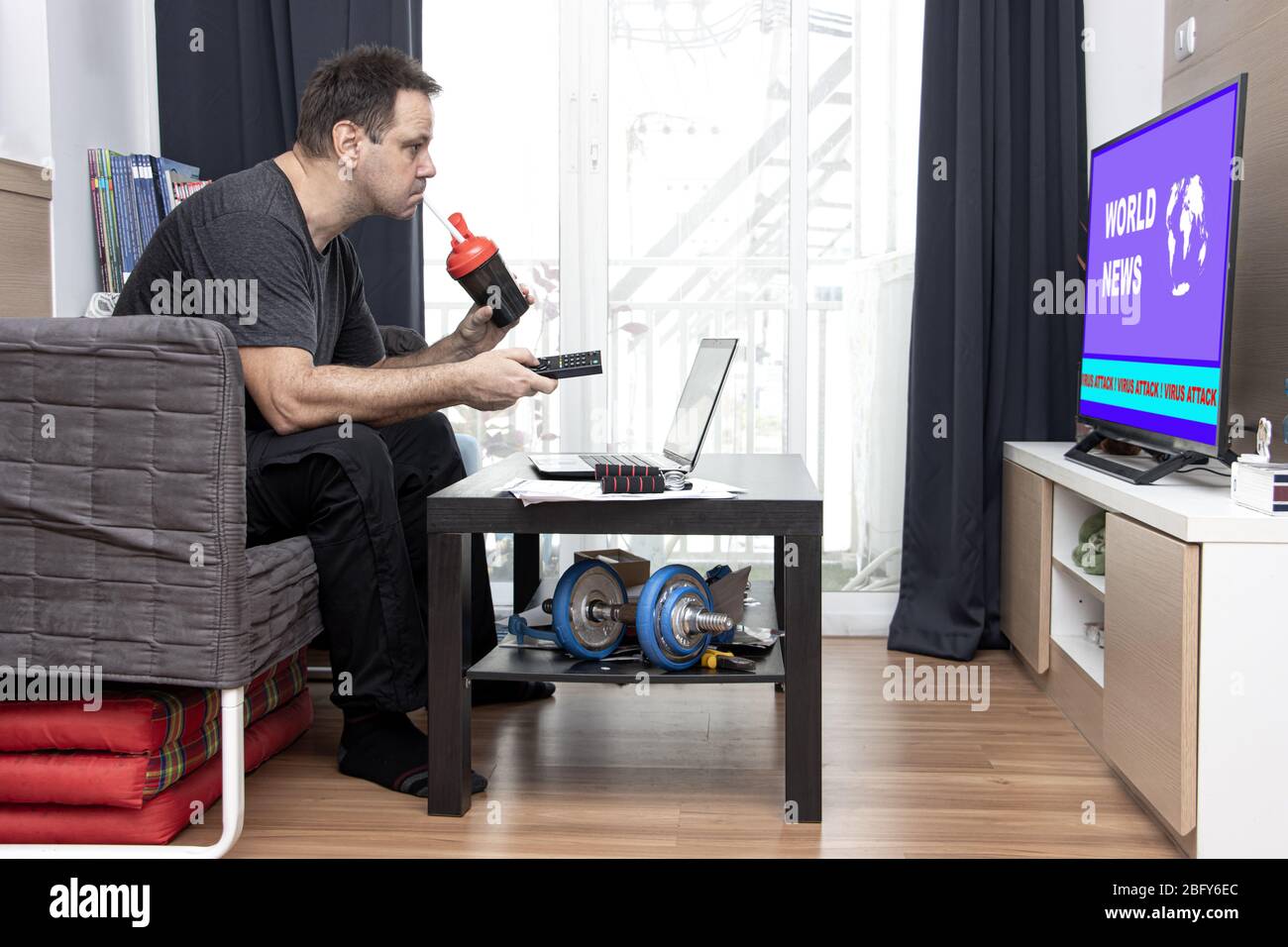 Un uomo guarda la televisione con le notizie TV circa il virus globale mentre si siede in soggiorno con il laptop su un tavolo. Ufficio domestico. Foto Stock