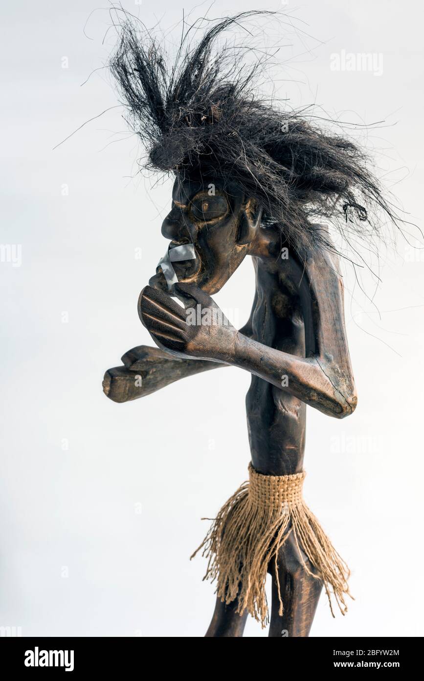 Figura intagliata in legno di uomo primitivo che indossa gonna di paglia e lunghi capelli neri a forma di cuce e nastrati su mouth.Concept; mobilità sociale, ignorata, silenziata. Foto Stock