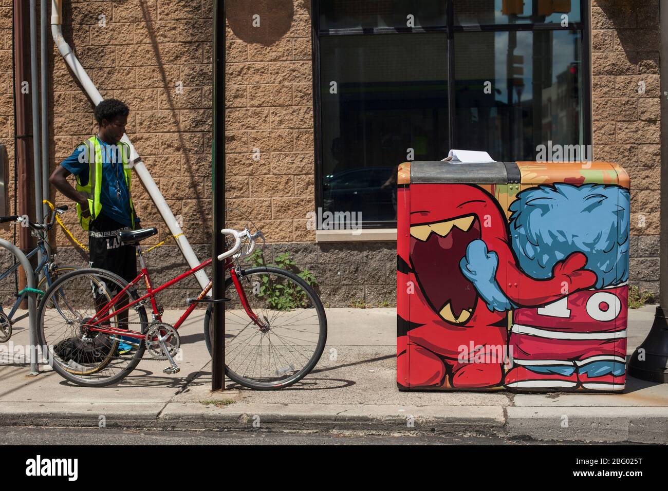 Giovane uomo nero con una bicicletta vicino a un cestino di carta da rifiuti decorato a South St, Philadelphia, Pennsylvania, USA – 4 agosto 2016: Foto Stock