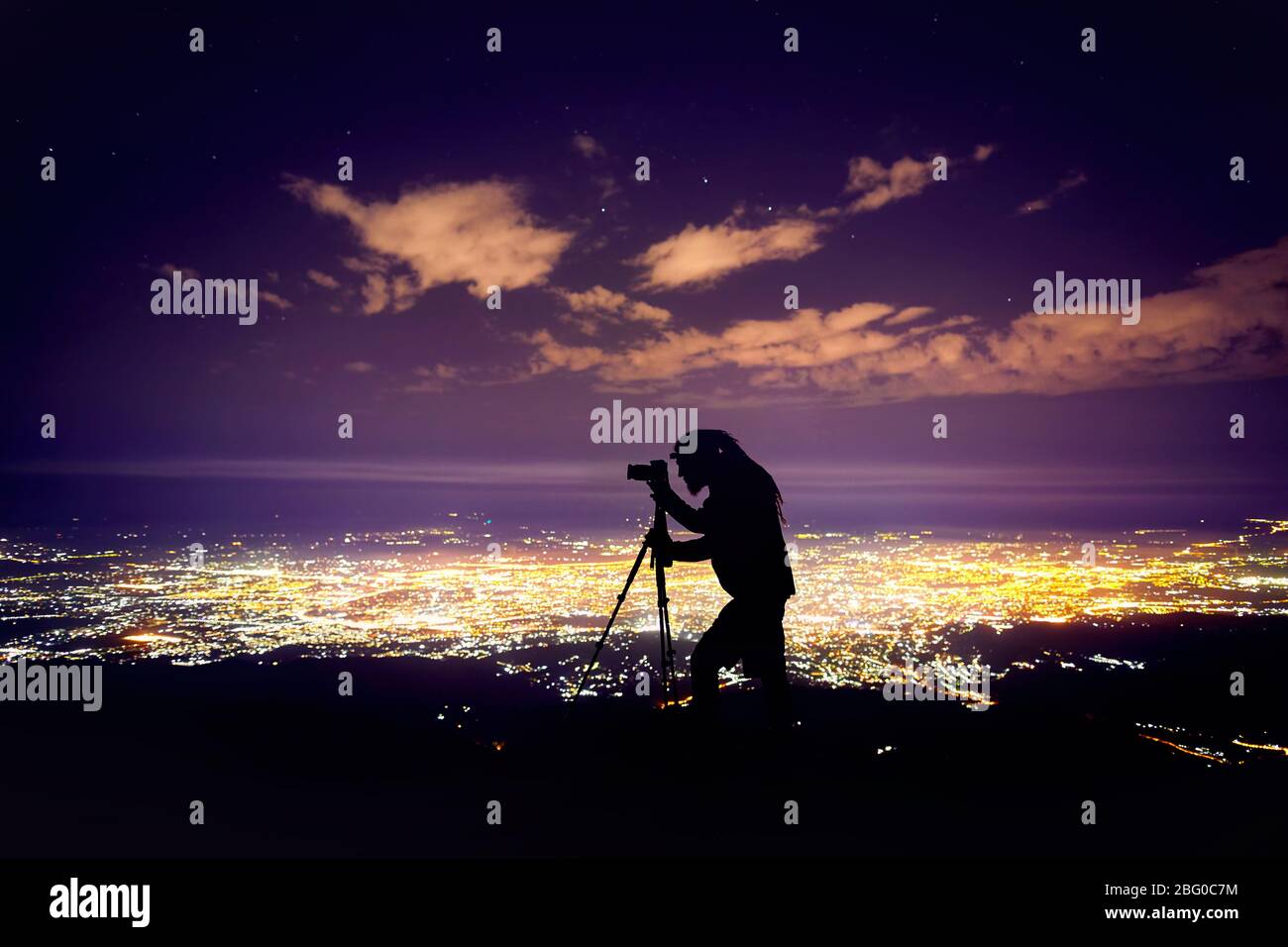 Fotografo con dreadlocks in silhouette tenendo girato con la fotocamera sul cavalletto contro il cielo notturno con stelle e incandescente città sfondo luminoso. Foto Stock