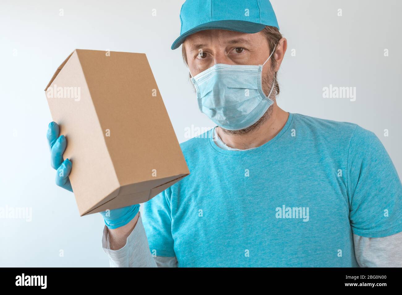 Consegna uomo che posa con pacchetto in indumenti protettivi che indossano cappello da baseball, maschera protettiva e guanti durante infezione virale pandemic outbre Foto Stock