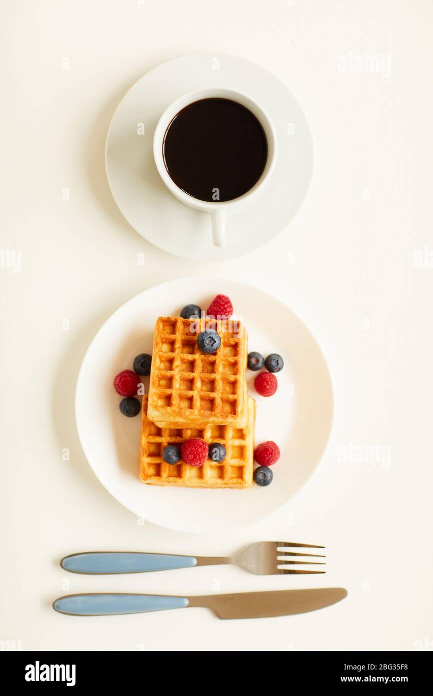 Vista dall'alto con una composizione minima di waffle dolci con aggiunta di frutti di bosco accanto a una tazza di caffè nero su sfondo bianco, spazio per la copia Foto Stock