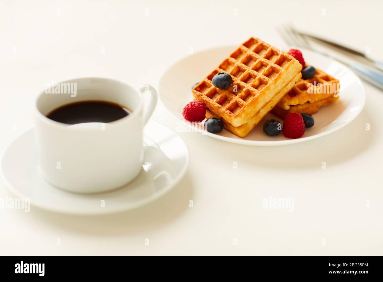 Primo piano con tonalità calde, waffle dolci da dessert con aggiunta di frutti di bosco accanto a una tazza di caffè nero su sfondo bianco, spazio per le copie Foto Stock