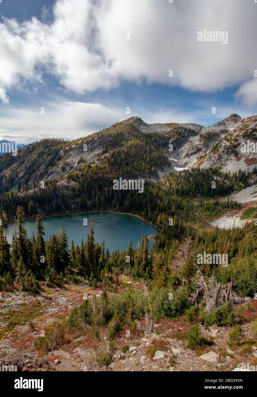 Un lago di montagna in un bacino è visibile dal lato di una montagna, circondato da larici occidentali, sotto un cielo blu con nuvola bianca e grigio chiaro Foto Stock