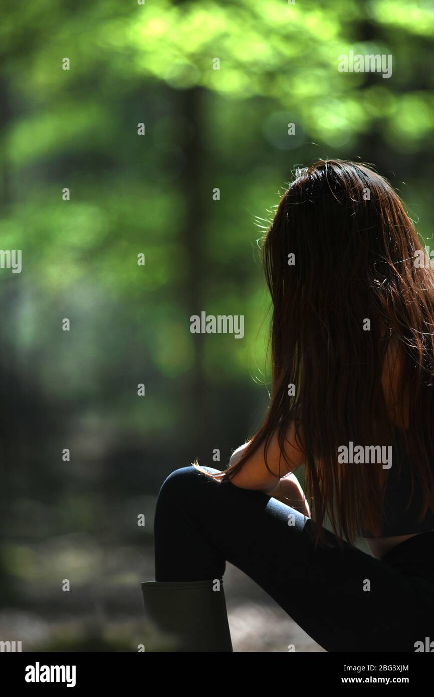 giovane ragazza pensierosa sedette in una luminosa e colorata giornata di sole nella foresta con il sole che illumina la nuova vita negli alberi, sfondo naturale per t Foto Stock