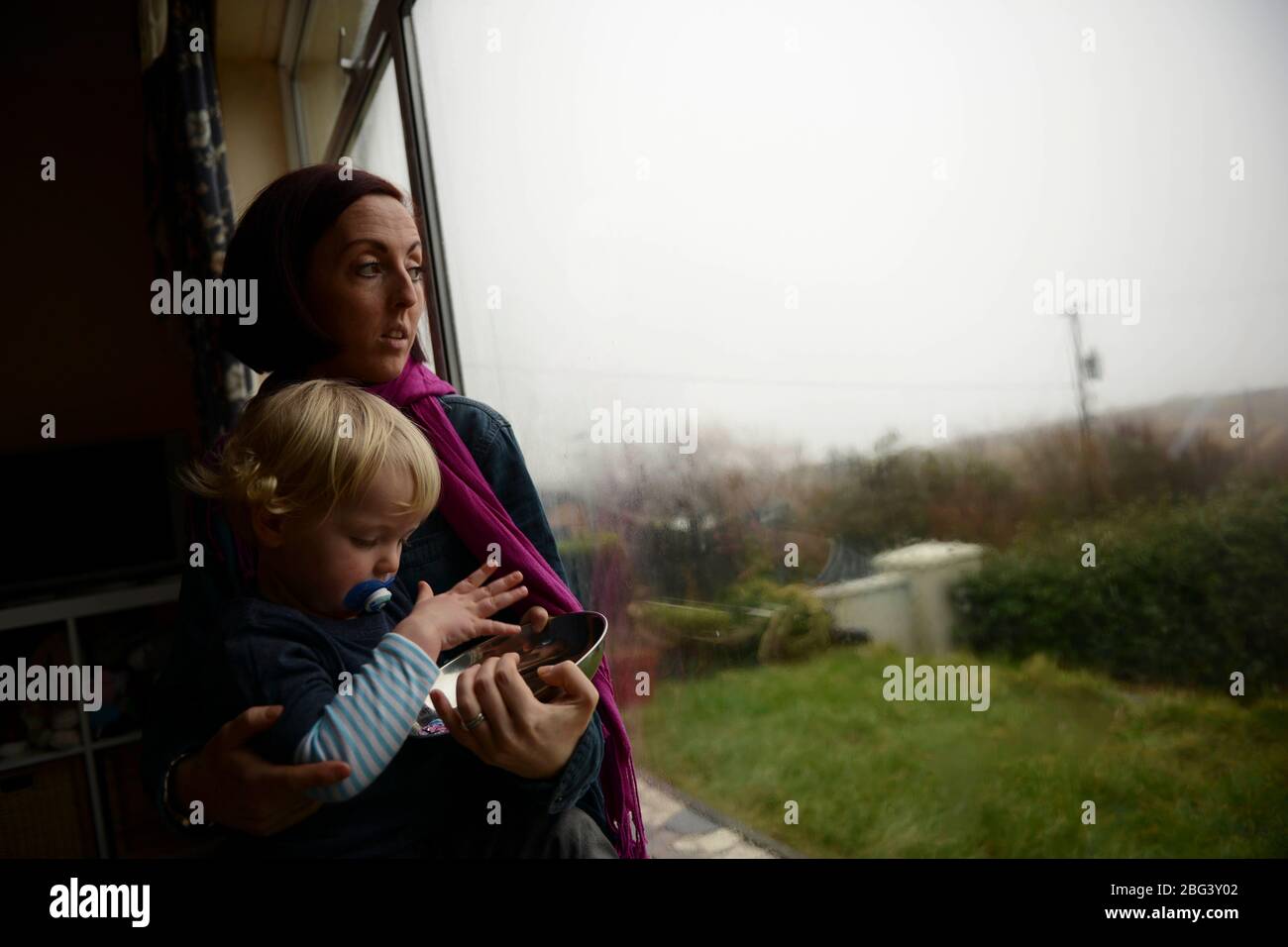Una giovane madre che soffre di depressione post-natale siede ad una finestra con il suo bambino Foto Stock