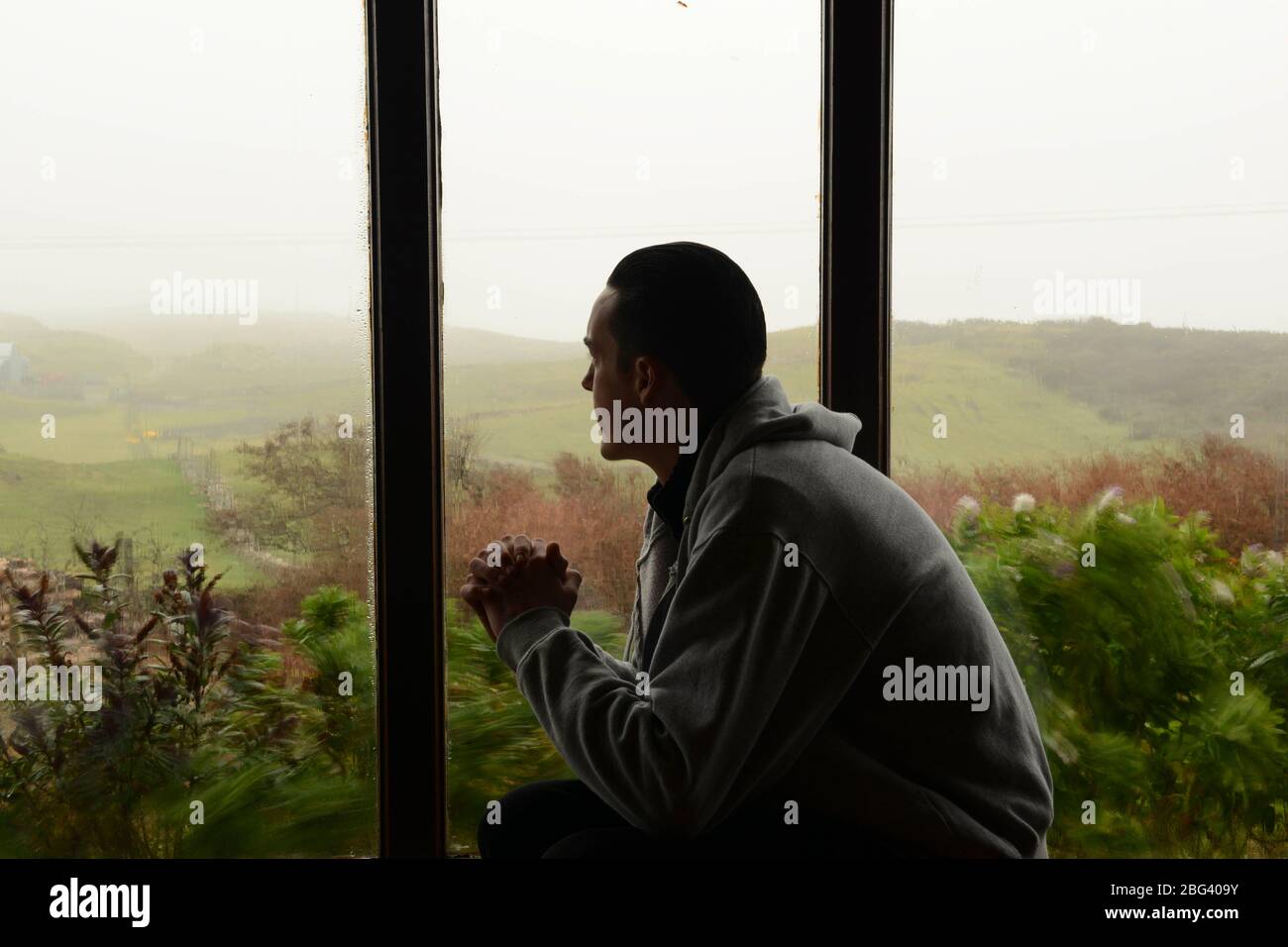 Un giovane uomo che soffre di depressione siede da una finestra che si chiede come passare la giornata avanti Foto Stock