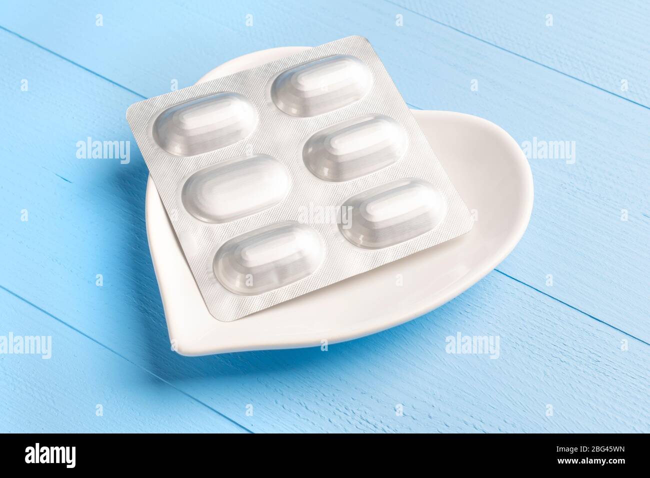 Blister di pillole mediche in un piatto bianco a forma di cuore su un blu di legno. Concetto di trattamento delle malattie cardiovascolari Foto Stock
