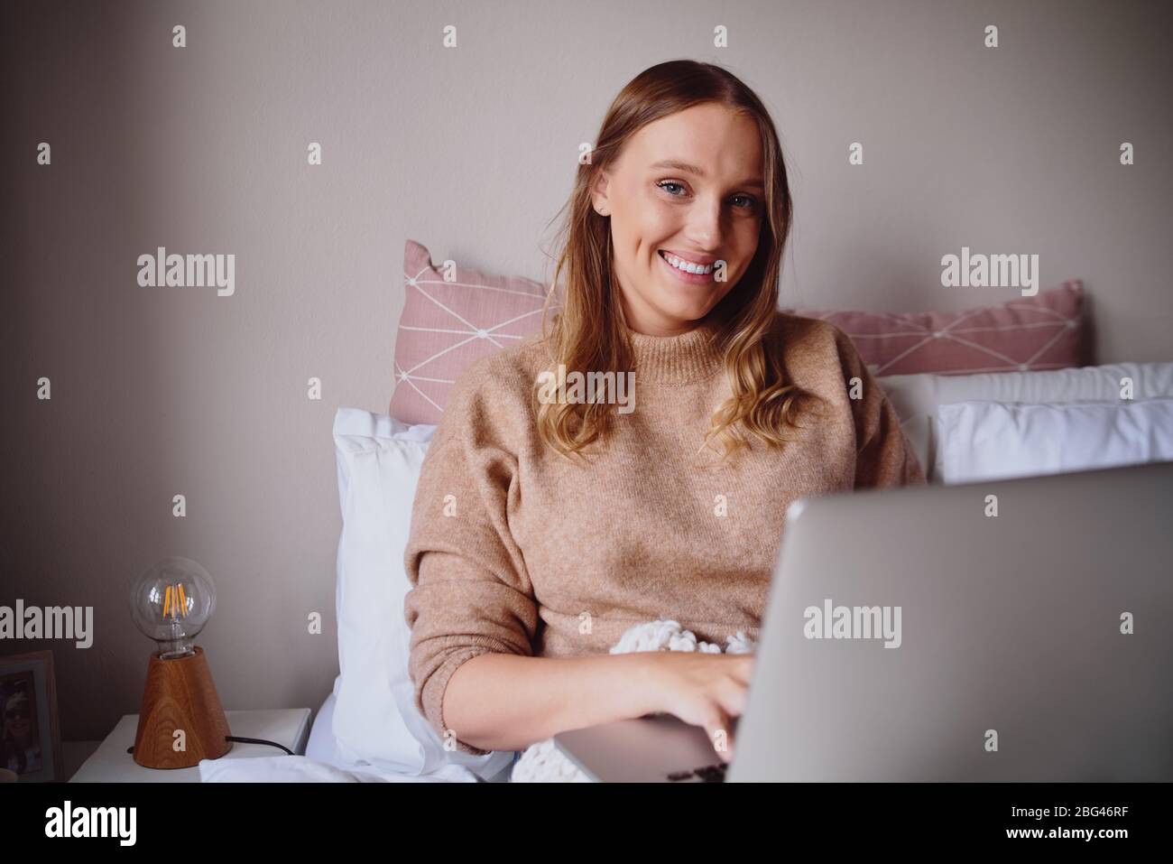 Felice freelancer femmina sedersi sul letto moderno e di scrivere laptop durante il periodo di blocco del virus corona sorridendo alla macchina fotografica Foto Stock