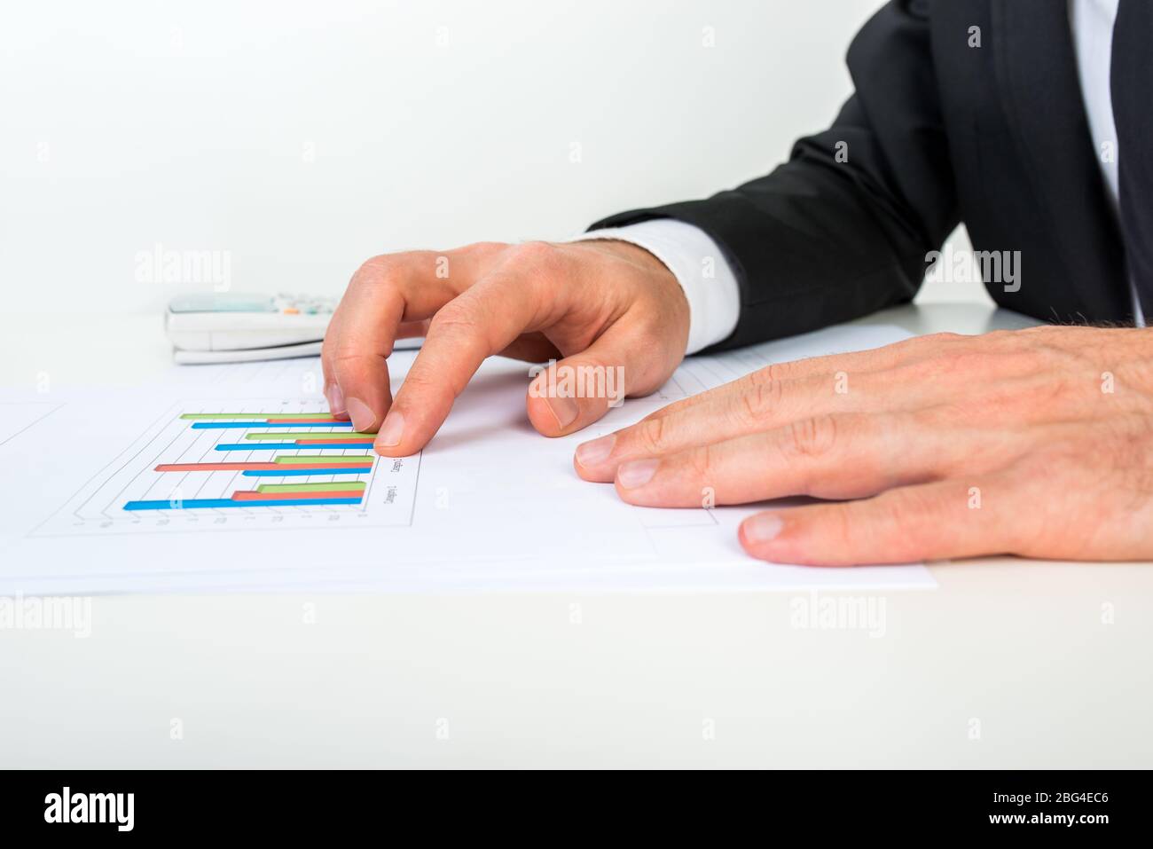 Vista laterale dell'uomo d'affari che analizza una serie di grafici a barre mentre si siede al suo banco bianco che punta ad un grafico a colonne, in un'analisi e strategia di affari Foto Stock