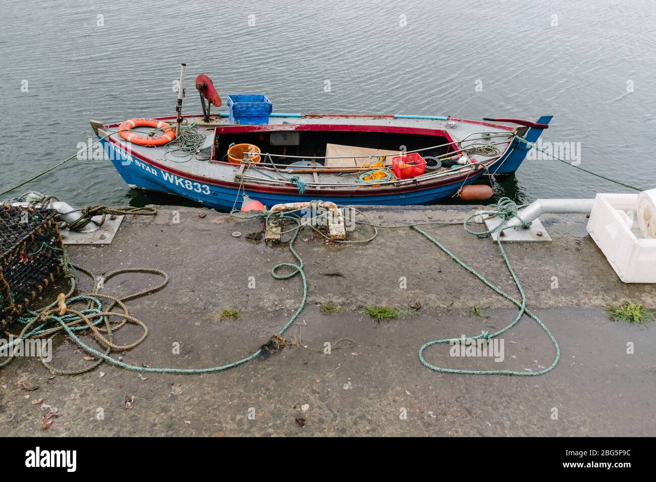 Piccola barca per la pesca dell'aragosta attraccata sul molo accanto alle creelle e alle scatole di pesce a Wick Harbour, Caithness, Scozia, Regno Unito. Foto Stock