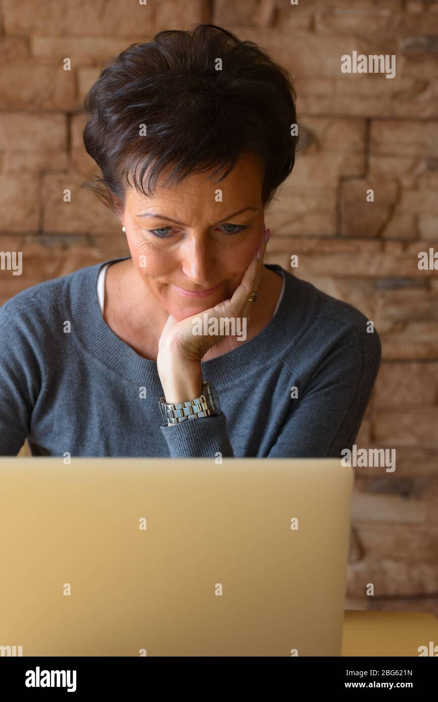 Donna matura bella poggiare mento a portata di mano mentre si utilizza il computer portatile su tavolo di legno contro parete di mattoni Foto Stock