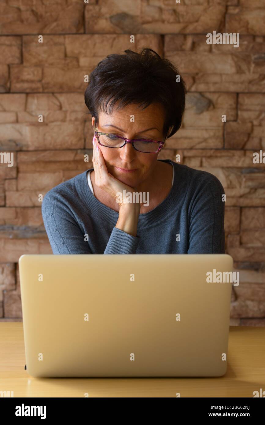 Donna matura e bella che usa il laptop mentre indossa gli occhiali e appoggia il mento a portata di mano Foto Stock
