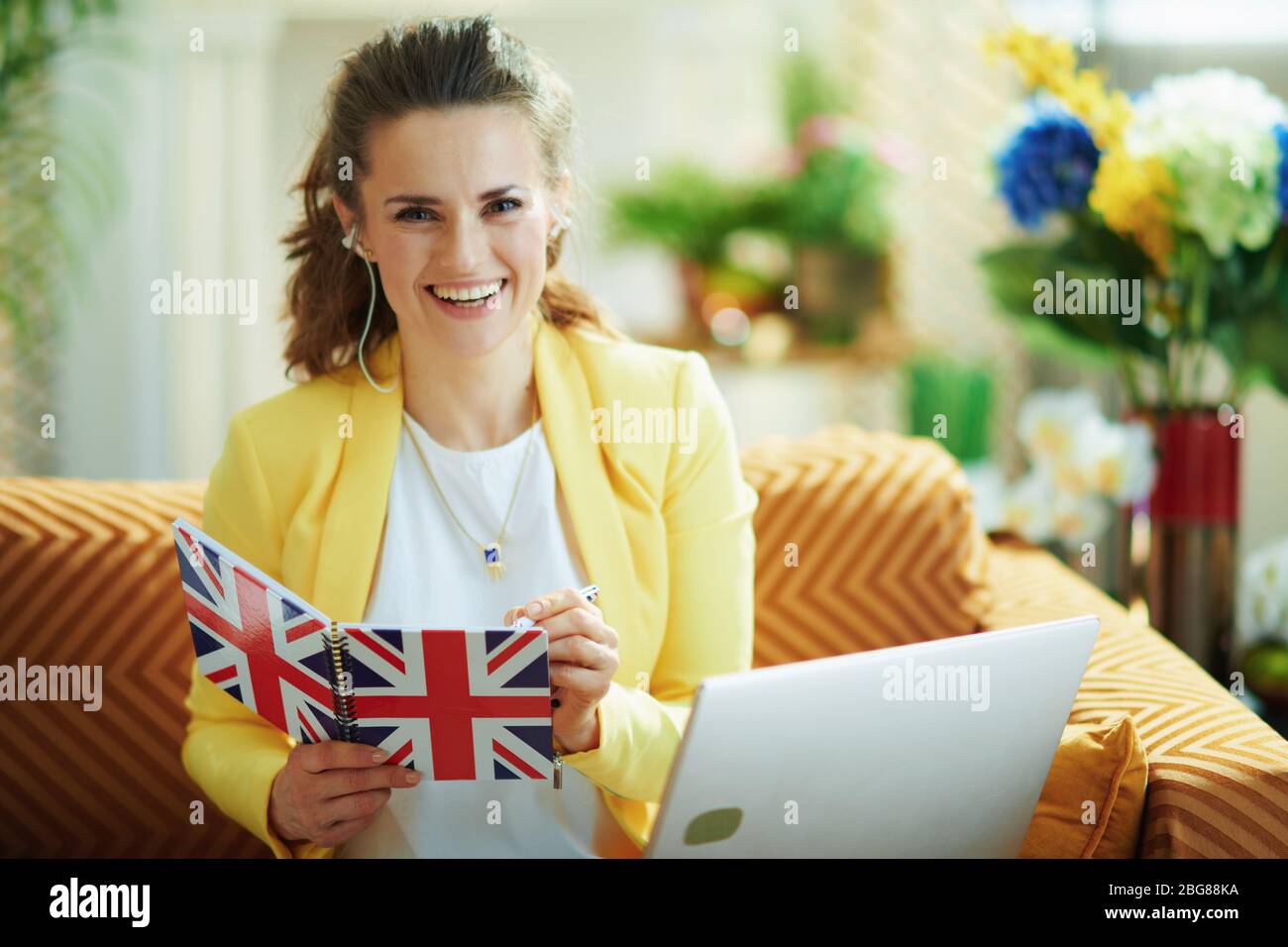 Sorridente elegante casalinga di mezza età in jeans e giacca gialla con notebook prendere appunti con una penna in un notebook bandiera del Regno Unito a casa moderna in giorno di sole. Foto Stock