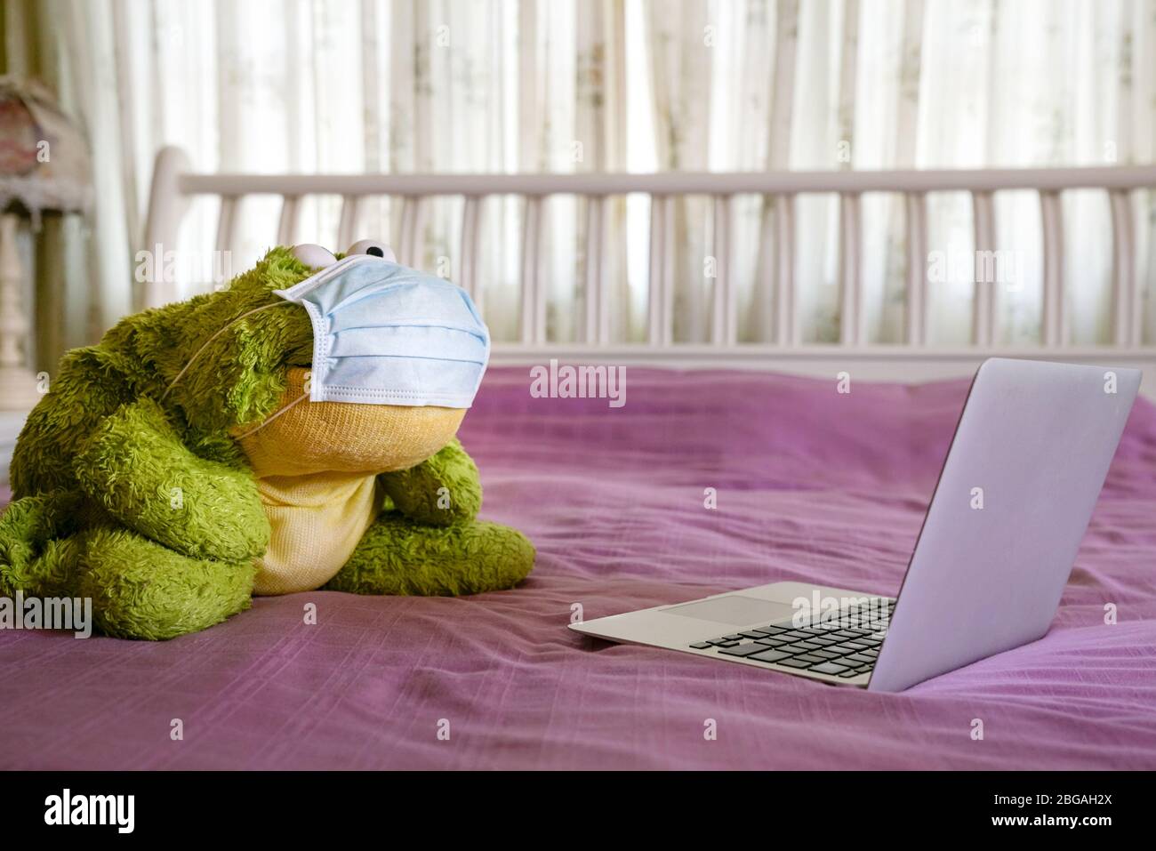 Giocattolo verde morbido con maschera medica e vista dello schermo del computer sulla parte superiore del letto. Lavoro a casa. Foto Stock
