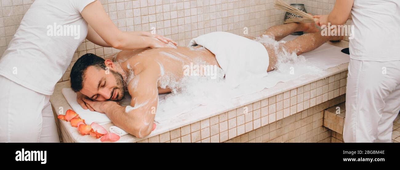 Uomo orientale che ottiene un massaggio delle gambe con una ginestra di bambù in una spa. Operatore hammam che fa massaggio in schiuma su una tavola calda in marmo di bagno orientale Foto Stock