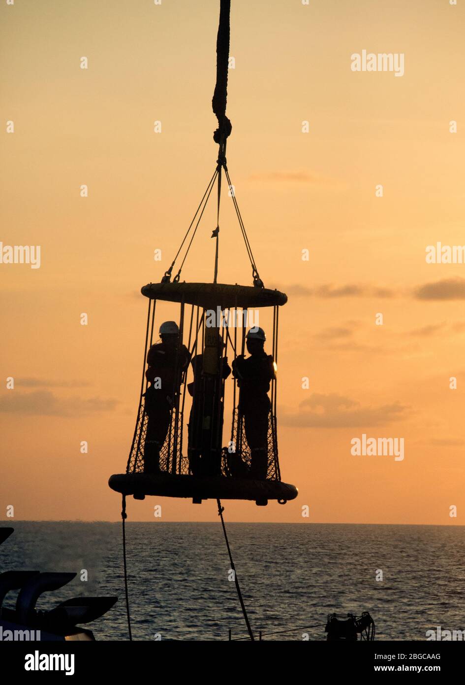 silhouette offshore operaio all'interno di un cestino personale impiccato in aria durante il trasferimento personale dalla chiatta da lavoro alla piattaforma petrolifera Foto Stock