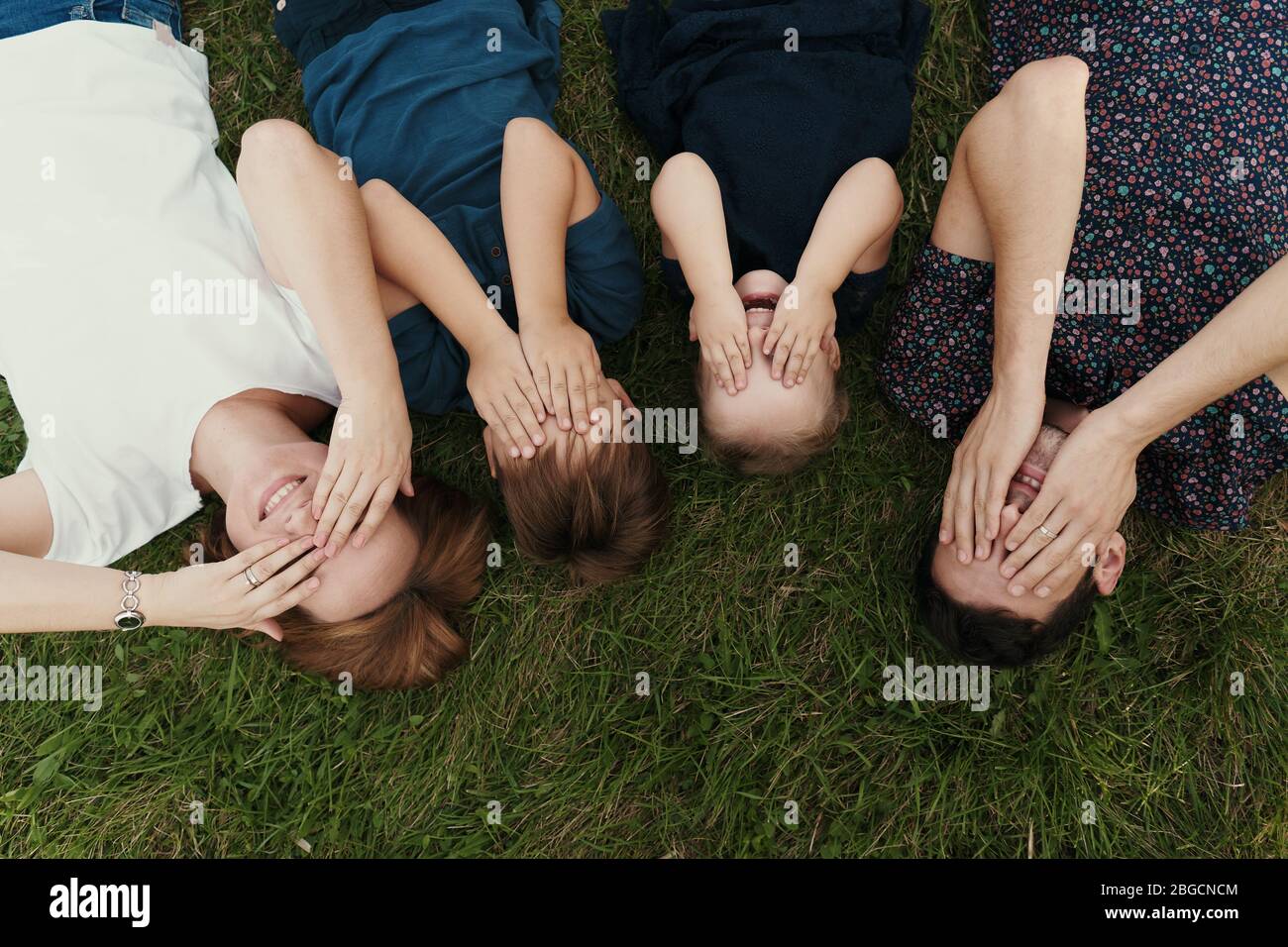 Giovane famiglia di quattro membri che giace sull'erba coprire gli occhi con le mani, creare ricordi per una vita Foto Stock