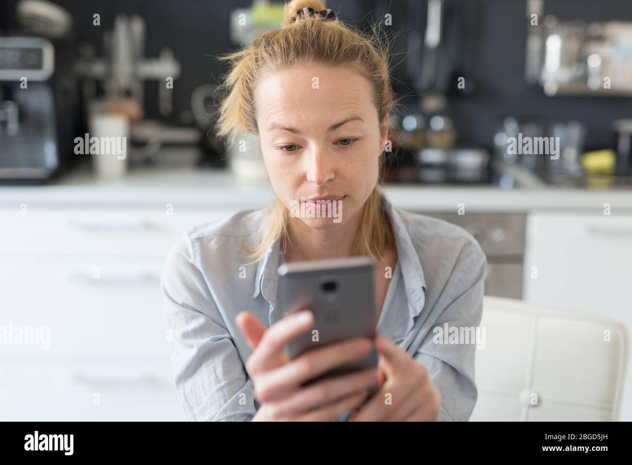 Donna preoccupata in casa in cucina utilizzando le app social media sul telefono per le chat video e le foto in contatto con i suoi cari. Rimani a casa Foto Stock