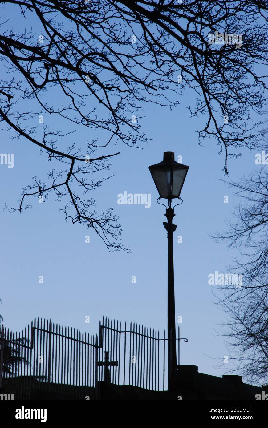 Luce da strada singola con recinzione sfalsata in ferro battuto e croce dietro con albero senza foglie curvare intorno alla luce tutto in silhouette Foto Stock