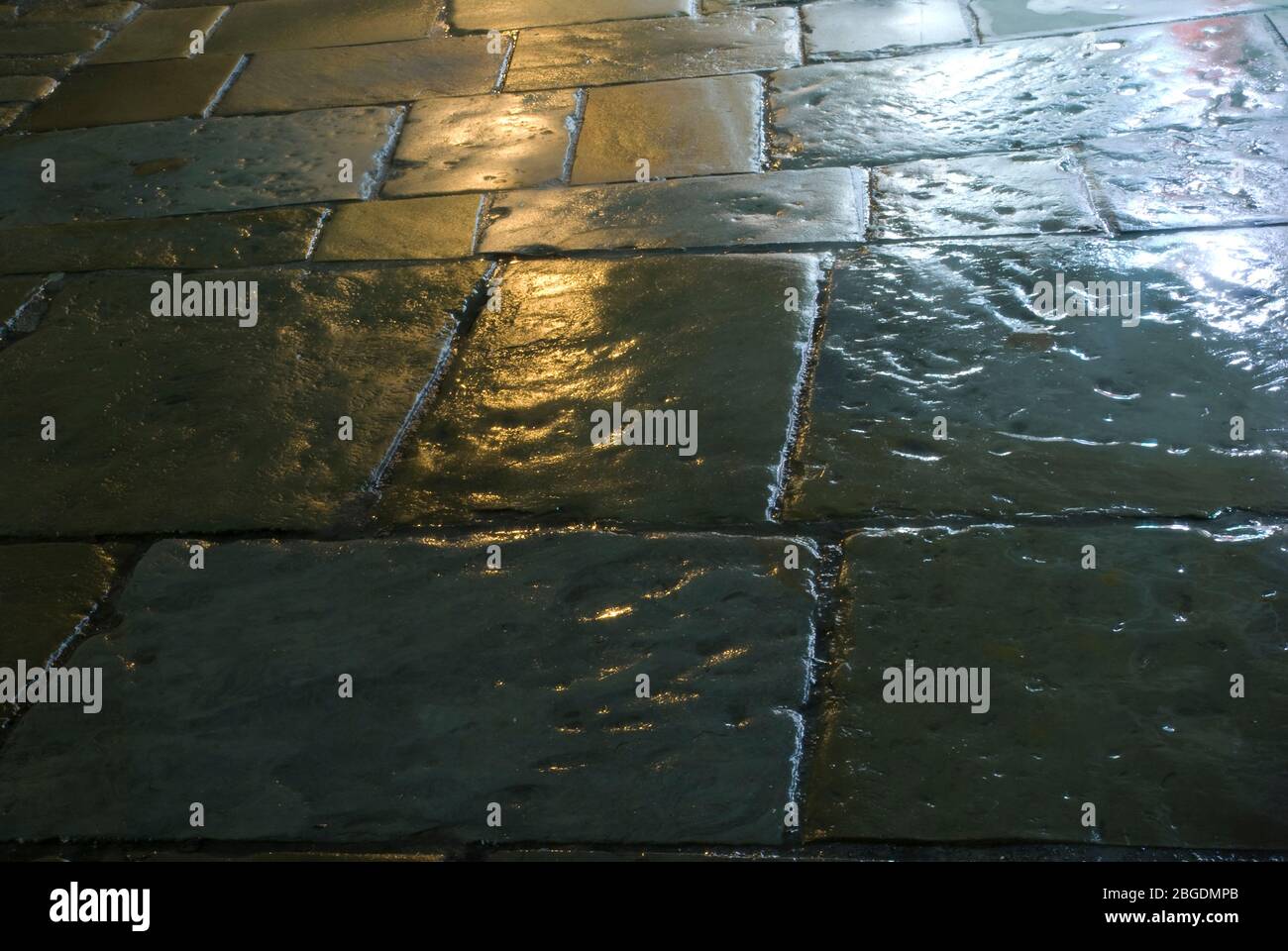 Pavimentazione in pietra di bandiera bagnata di notte che mostra le pietre di bandiera in primo piano e riflessi di illuminazione stradale sulla superficie bagnata Foto Stock