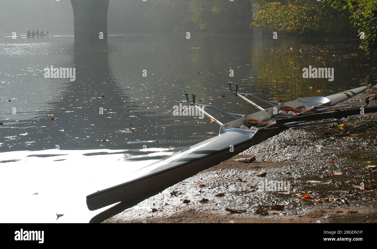 Riverside scena con barca da corsa per due legate mentre un quattro e cox barca passare sotto gli archi di un ponte in una giornata di autunno misteriosa Foto Stock