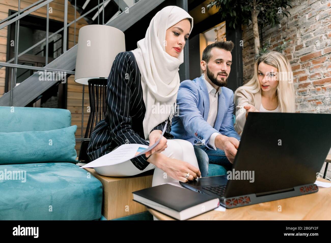 Tre giovani colleghi multietnici, uomo e donna caucasica, signora musulmana in hijab, che hanno conversazione e uso di laptop, seduti su divano blu Foto Stock