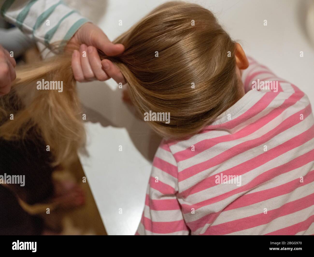 Una ragazza di quattro anni con i capelli biondi lunghi sta avendo i suoi capelli spazzolati dalla sua mamma dopo un lavaggio. Sta guardando via verso una televisione (fuori dal pic Foto Stock