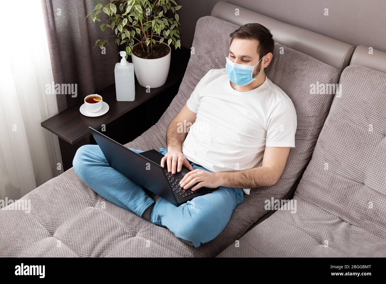 Luogo di lavoro domestico. Uomo freelancer in faccia maschera chirurgica che lavora da casa utilizzando il laptop. Comodo ufficio sul divano. Coronavirus covid-19 distanza sociale Foto Stock