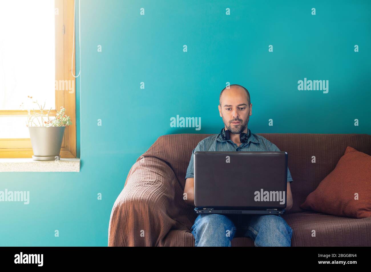 Uomo seduto su un divano che lavora da casa con il suo laptop e le cuffie Foto Stock