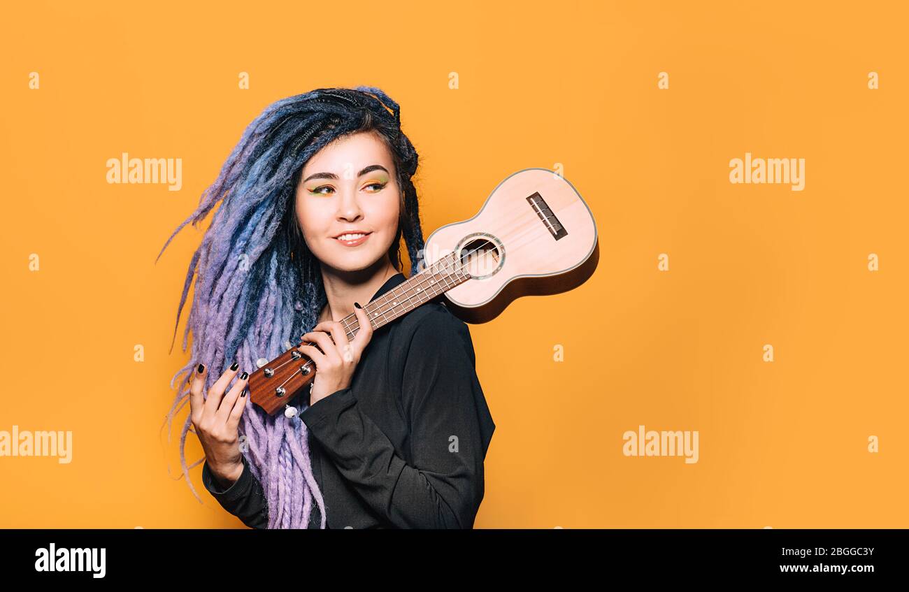 ritratto di donna hipster con i treadlocks viola e ukulele nelle sue mani su uno sfondo arancione. Strumento musicale ukulele Foto Stock