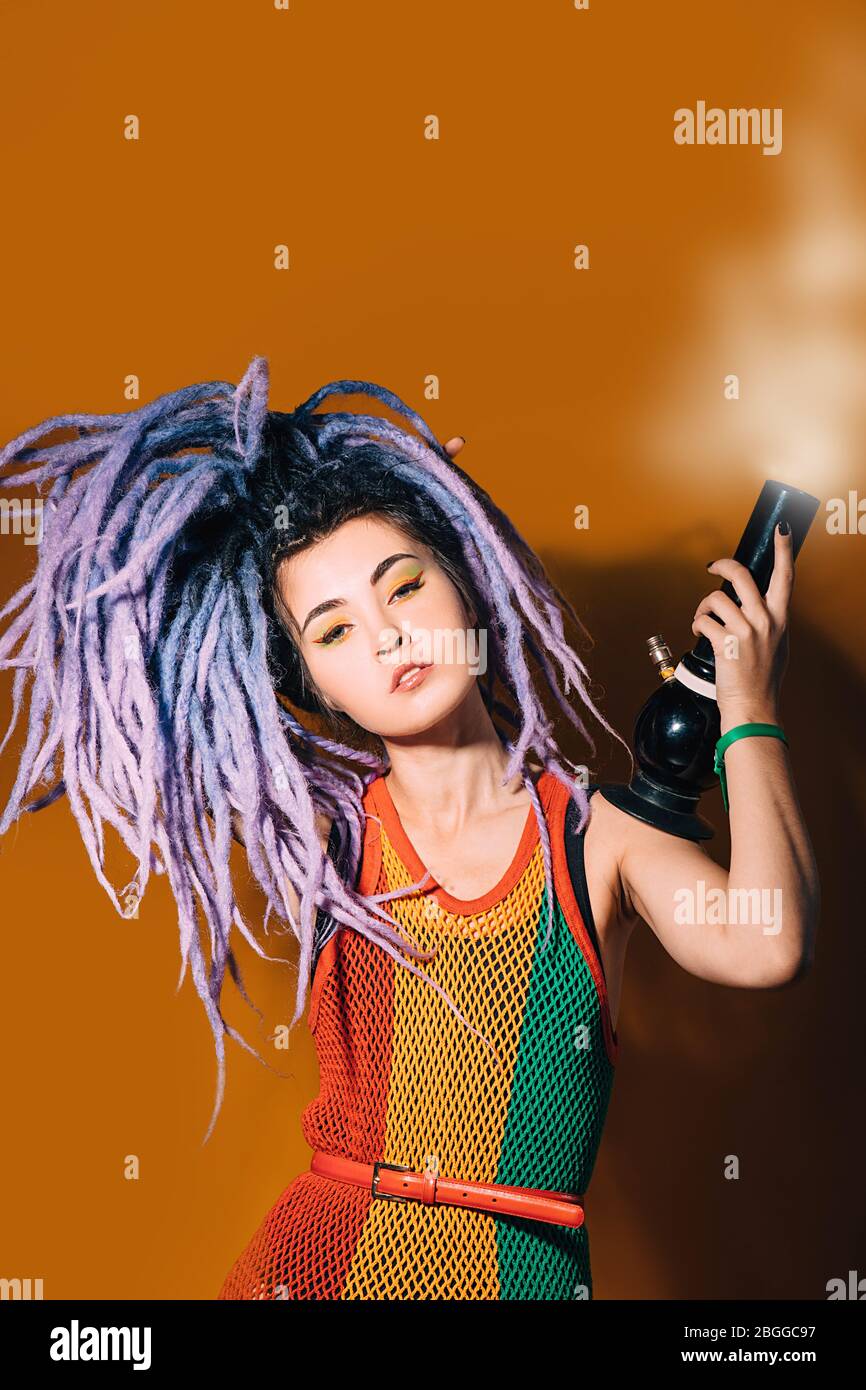 Donna Rastafariana fuma marijuana. Cannabis fumatore bong. Donna in abito rasta e lucchetti viola su sfondo arancione Foto Stock
