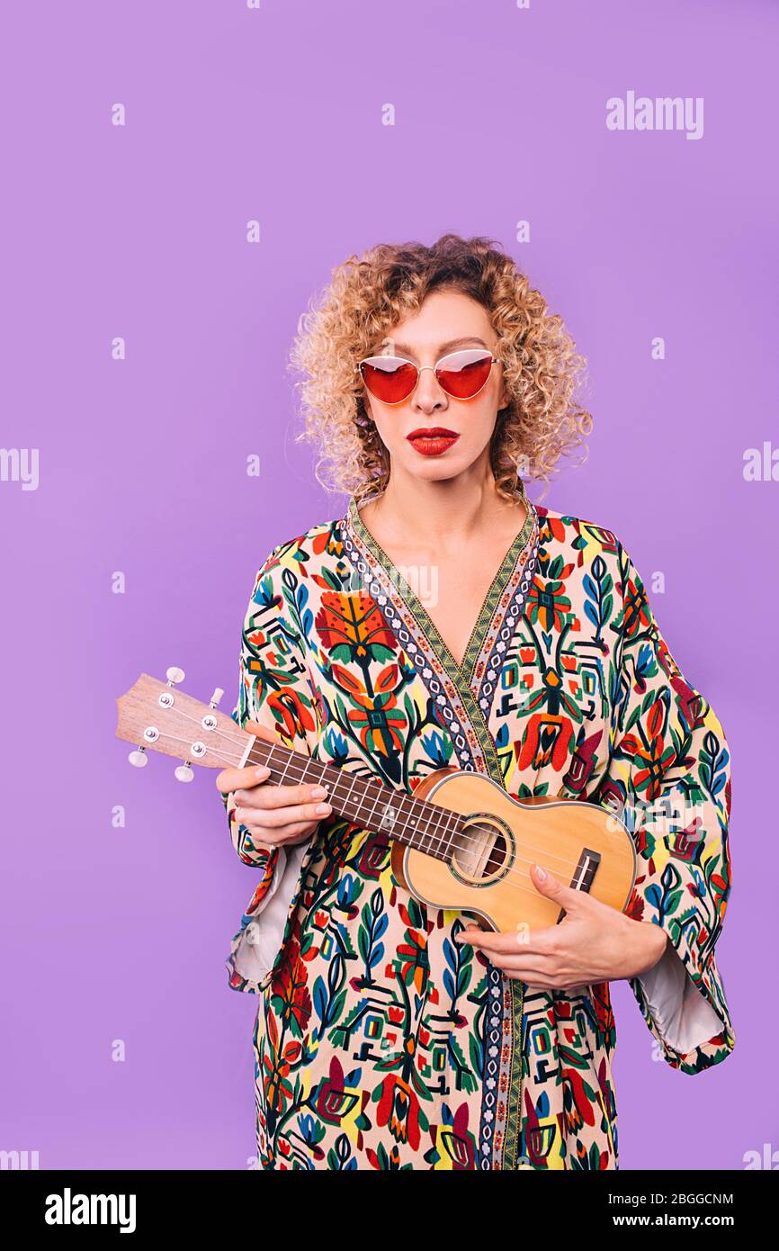 donna hipster in occhiali rosa e un bel vestito che tiene uno strumento musicale ukulele ukulele su sfondo viola Foto Stock