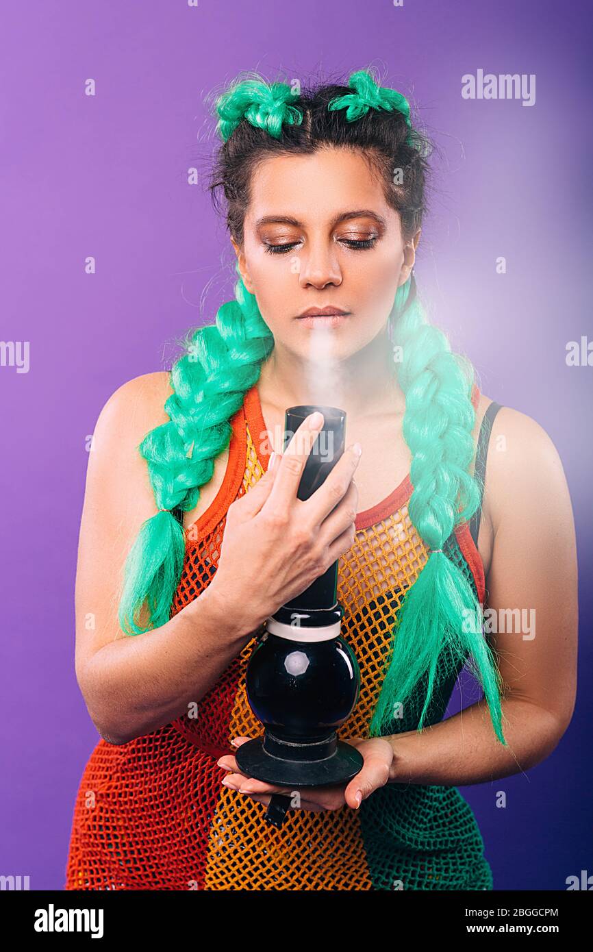Grande dongolo di vetro nelle mani di una donna hipster. La donna fuma cannabis. Pettinatura femminile con kanekalon verde su sfondo viola Foto Stock