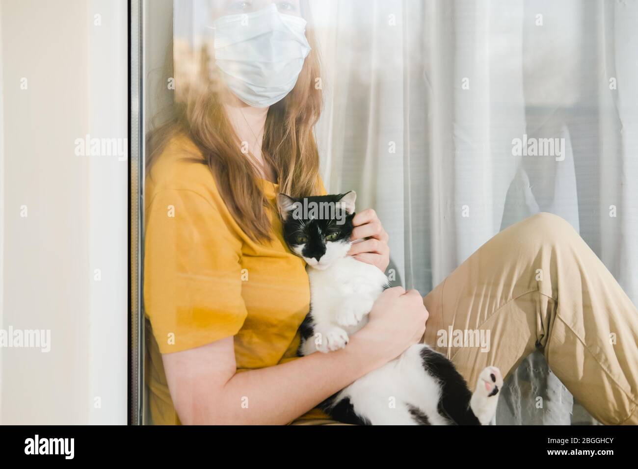 Triste ragazza in maschera medica protettiva e maglietta gialla seduta sul davanzale con un gatto Tuxedo nero e bianco durante una corona di quarantena pandemica Foto Stock