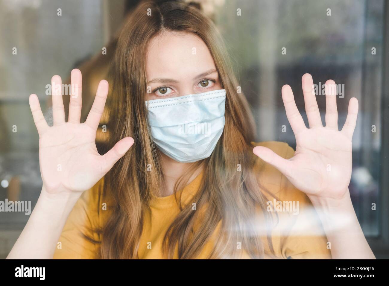 Triste ragazza in maschera medica protettiva e maglietta gialla in piedi a casa sua e guarda fuori la finestra. Covid-19 coronavirus pandemico. Isolamento a hom Foto Stock