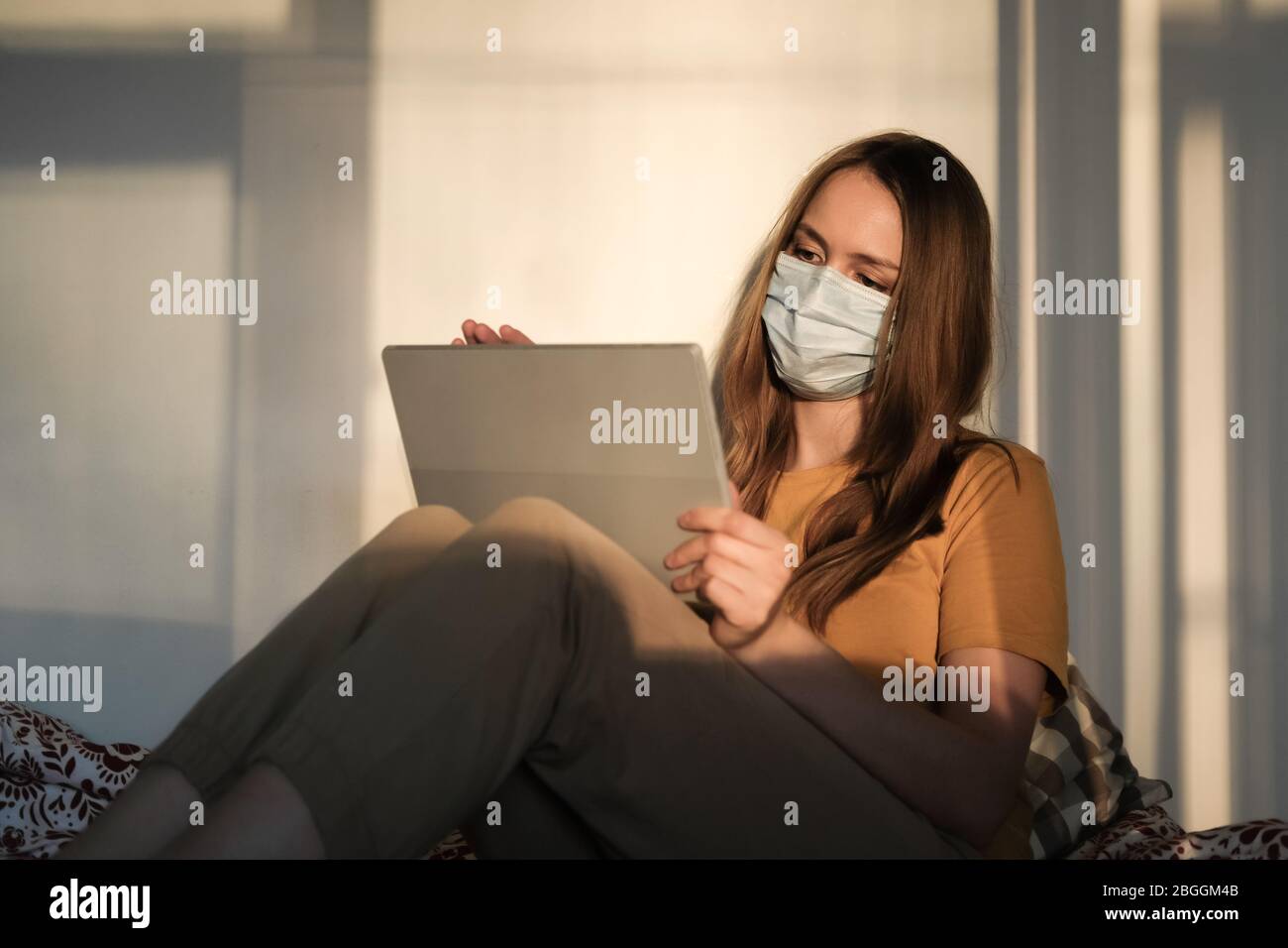 Giovane ragazza in maschera medica protettiva e T-shirt gialla sta studiando o lavorando sul letto sulla compressa. Covid-19 quarantena coronavirus pandemico. Istruzione Foto Stock